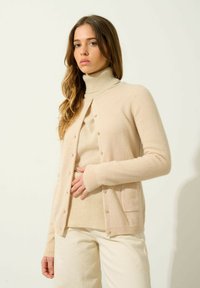 Cárdigan de lana beige con mangas largas, cuello redondo, botones frontales y un bolsillo. Combinado con un jersey de cuello alto a juego y pantalones de color crema.
