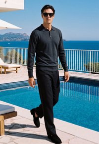 Homme portant des lunettes de soleil noires et des vêtements sombres marchant près d'une piscine avec vue sur la mer et des montagnes en arrière-plan par une journée ensoleillée.