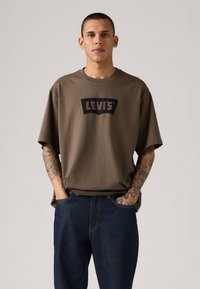 Braunes Baumwoll-T-Shirt in Übergröße mit kurzen Ärmeln und einem schwarzen Levi's-Logo-Graphic. Getragen mit dunkelblauen Denim-Jeans.