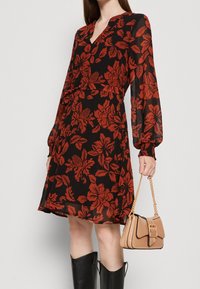 Robe noire à fleurs avec des fleurs rouges, manches longues, décolleté en V et poignets froncés. Associée à un sac à main beige avec une bandoulière chaîne et un détail de boucle.