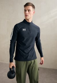 Fekete, hosszú ujjú sportpóló cipzáros gallérral, amelyen egy fehér Under Armour logó található a mellkason, zöld cargo nadrággal párosítva, és egy kettlebellt tart.