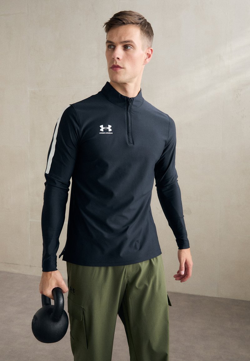 Fekete, hosszú ujjú sportpóló cipzáros gallérral, amelyen egy fehér Under Armour logó található a mellkason, zöld cargo nadrággal párosítva, és egy kettlebellt tart.