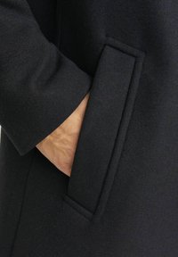 Palton negru din lână, cu un buzunar lateral, având o textură netedă și un design curat, structurat. Vedere de aproape care evidențiază detaliile țesăturii și buzunarului.