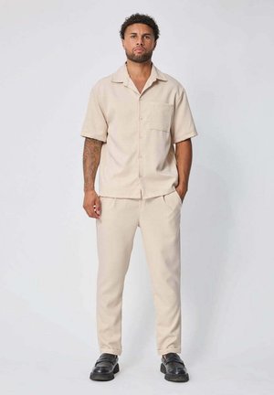 Homme aux cheveux bouclés et à la barbe portant une chemise beige à manches courtes boutonnée et un pantalon assorti, debout, les mains dans les poches et des chaussures noires.
