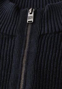 Pull en maille côtelée bleu marine avec une fermeture éclair argentée dotée d'un tirant de marque. Tissu texturé à rayures verticales, ajoutant de la profondeur.