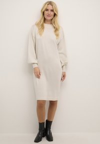 Strickkleid mit langen Ärmeln in hellbeige, lockerer Schnitt, gerippten Bündchen und runder Halslinie. Getragen mit schwarzen Stiefeletten bis zum Knöchel.