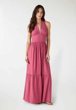 Donna con i capelli lunghi che indossa un vestito rosa senza maniche, con scollo a V profondo e lunghezza fino alle caviglie, e sandali, in piedi davanti a uno sfondo bianco uniforme.