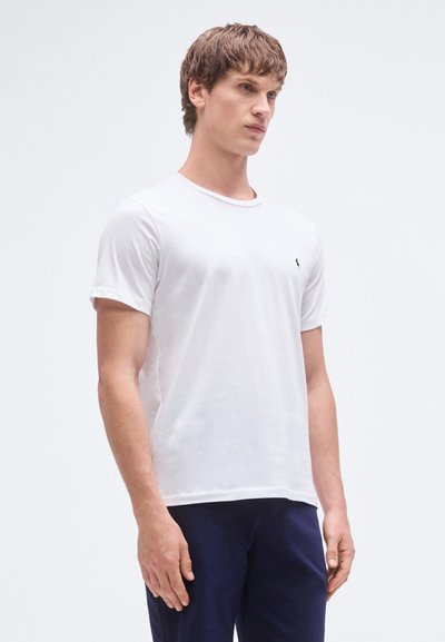 T-shirt branca de algodão com mangas curtas, gola redonda e um pequeno logótipo escuro no peito. O modelo está a usar calças azul-marinho.