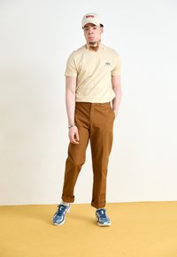 Beige póló logóval, barna szabott nadrág, és kék sportcipő. Fehér sapka piros felirattal. Egyszerű dizájn, tompított színek, laza öltözet.