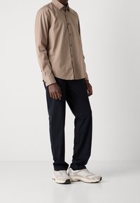 Hombre con una camisa de botones de manga larga beige, pantalones negros y zapatillas blancas y grises, de pie contra un fondo blanco liso.