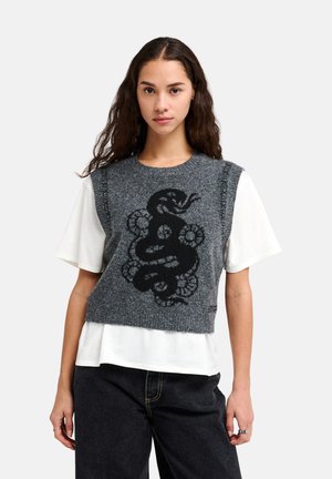 Jonge vrouw met lang krullend haar draagt een grijs mouwloos gebreid vest met een zwart slang- en bloemenmotief over een wit shirt met korte mouwen.