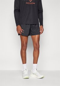 T-shirt de sport noir à longues manches avec logo orange ; shorts noirs avec texture à motif et petit graphique orange ; chaussures de course blanches avec accents verts.