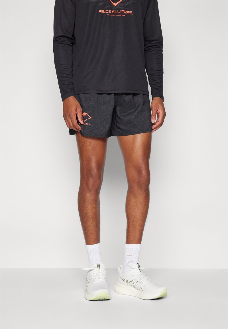 T-shirt de sport noir à longues manches avec logo orange ; shorts noirs avec texture à motif et petit graphique orange ; chaussures de course blanches avec accents verts.