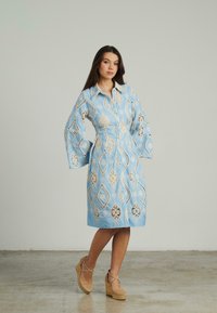 Blaues Hemdkleid mit weißem Blumenmuster und Spitzenverzierungen, mit langen Glockenärmeln, tailliertem Schnitt und knielangem Saum. Getragen mit Wedge-Sandalen.