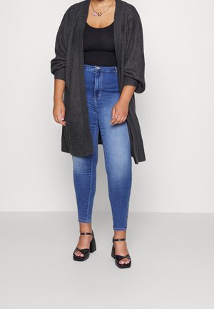 Femme portant un cardigan gris foncé ouvert sur le devant, un débardeur noir, un jean skinny bleu, des sandales à talons noires et un collier en or avec un pendentif floral.