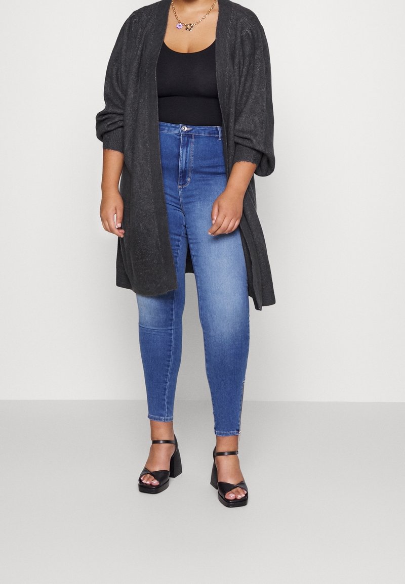 Femme portant un cardigan gris foncé ouvert sur le devant, un débardeur noir, un jean skinny bleu, des sandales à talons noires et un collier en or avec un pendentif floral.
