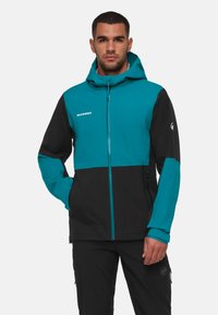 Tarnblauer und schwarzer Jacke mit einem Reißverschluss vorne, angenommener Kapuze und Logo auf der Brust. Sie zeichnet sich durch ein schlankes, tailliertes Design mit kontrastierenden Farben aus.