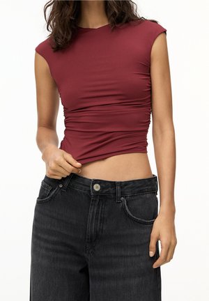 Femme portant un haut sans manches froncé de couleur bordeaux et un jean noir taille haute, avec une main levant légèrement le haut pour révéler la taille.
