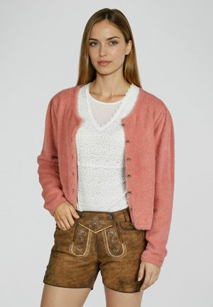 Femme portant un cardigan rose en peluche sur un haut en dentelle blanche et un short en cuir brun brodé, posant devant un fond uni.