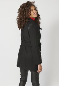 Cappotto di lana nero con vita a cintura, caratterizzato da dettagli in pelle sulle spalle. Indossato sopra leggings neri a trama.