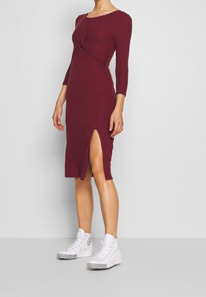 Kobieta ubrana w dopasowaną, długorękawową, bordową sukienkę midi z przednim rozcięciem, w połączeniu z białymi sneakersami za kostkę na szarym tle.
