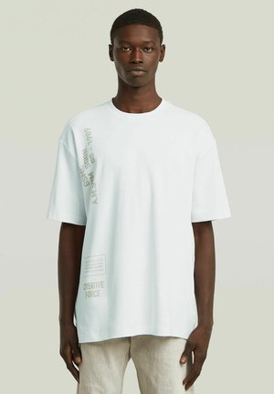 Ung mand med mørk hud iført en løs hvid T-shirt med beige tekst og lyse bukser, stående foran en ensfarvet baggrund.