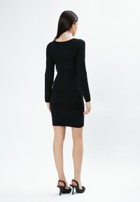Schwarzes, langärmliges Bodycon-Kleid mit strukturiertem Muster, auf dem "INFLUENCER" steht, mit rundem Rückenausschnitt und figurbetontem Schnitt, kombiniert mit schwarzen Absätzen.