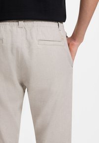 Pantalones casuales de lino beige con cinturilla elástica, usados por una persona con camisa negra, con la mano colocada en el bolsillo delantero derecho.
