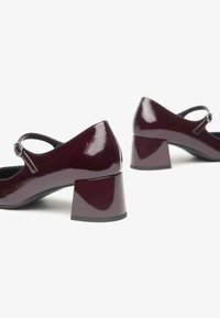 Dei tacchi in vernice bordeaux con punta rotonda, un dettaglio a fibbia sulla cinghia e un design con tacco a blocco. Texture liscia, finitura elegante.