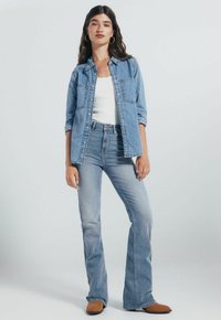 Denim overhemd met twee borstzakken, lichtblauwe kleur. Witte tanktop eronder. Gewijde lichtblauwe jeans, bruine enkellaarsjes, staande pose.