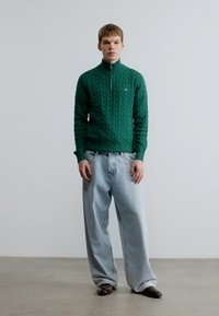 Zelený pletený sveter s vysokým golierom, v kombinácii s svetlomodrými džínsami s širokými nohavicami a tmavými topánkami, na modeli na jednoliatej pozadí.