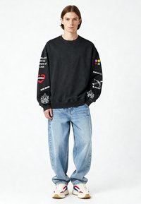 Felpa nera con grafiche stampate in rosso, bianco e colori sulle maniche. Abbinata a jeans blu larghi e sneakers bianche.