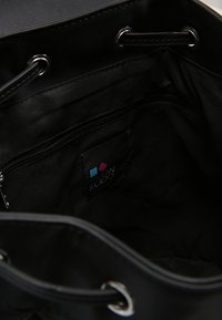 Intérieur d'un sac fourre-tout en tissu noir avec ferrures argentées, comprenant une poche zippée et une étiquette de marque en bleu et rouge. Texture lisse, design spacieux.