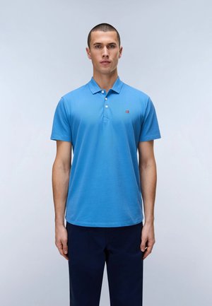 Jeune homme debout, face à l'avant, portant un polo bleu à manches courtes et un pantalon foncé sur un fond clair uni.