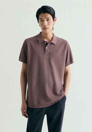 Młody mężczyzna z czarnymi włosami, ubrany w brązową koszulkę polo z krótkim rękawem i ciemne spodnie, stojący na tle jednolitego jasnego tła.