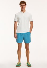 Biely polo tričko, svetlomodré kordové šortky, zelené tenisky so žltými šnúrkami. Model stojí s rukami v vreckách na neutrálnom pozadí.