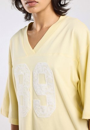 Person wearing a pale yellow V-neck shirt with large white lace-textured number "89" on the front, with short sleeves and loose fit.