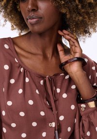 Brązowa bluzka w grochy z wiązaniem przy szyi, wykonana z lekkiego materiału. Dodatki to ciemne żywiczne bransoletki na nadgarstku.