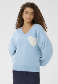 Pull bleu clair à col en V, fabriqué en matière douce et duveteuse ; possède un motif de cœur blanc sur la poitrine ainsi que des poignets et un ourlet côtelés.