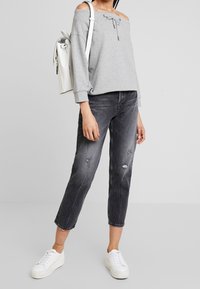 Sudadera gris de hombros caídos con cordón, combinada con jeans negros desgastados y zapatillas blancas. Mochila blanca colgada de un hombro.