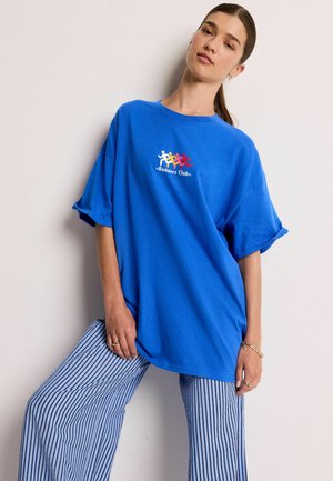 Kongelig blå oversized t-shirt med korte ærmer, prydet med et multifarvet "Runners Club" logo. Sammen med blå og hvide stribede bukser.