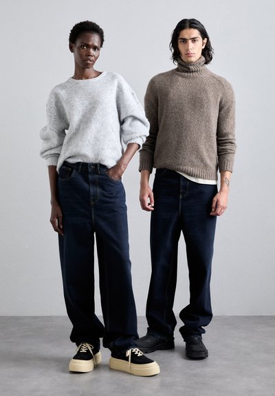 Two models stand side by side. The first wears a light gray, loose-knit sweater and high-waisted dark jeans. The second sports a brown, ribbed turtleneck and dark jeans. Both wear chunky sneakers.