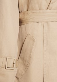 Trench-coat beige avec une ceinture à la taille, une boucle sur la manche et une poche en biais sur le côté.