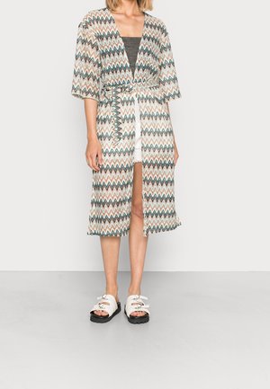 Femme portant un peignoir mi-long à motifs avec des rayures en zigzag, ceinturé à la taille, par-dessus un haut noir et un short blanc avec des sandales blanches.