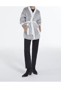 Person som bär svarta byxor, svarta stövlar och en vit och grå stickad cardigan med fickor och midjeband.