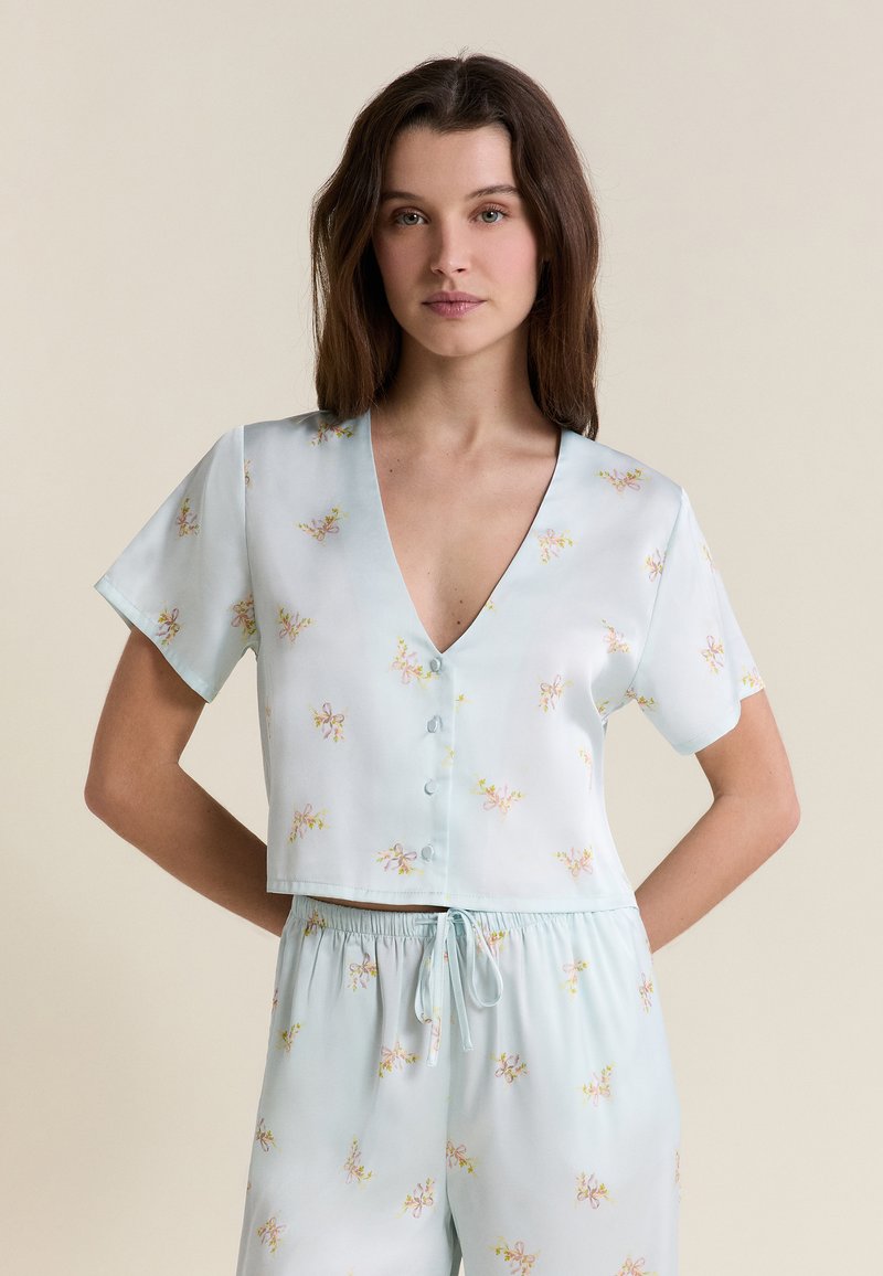 Joven mujer con blusa de manga corta azul claro con botones y pantalones a juego con cordón y pequeños estampados florales sobre fondo neutro.