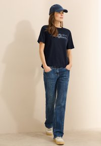 Camiseta navy con un estampado gráfico, combinada con unos pantalones vaqueros de corte recto de color azul claro y zapatillas blancas. Una gorra navy a juego completa el atuendo.