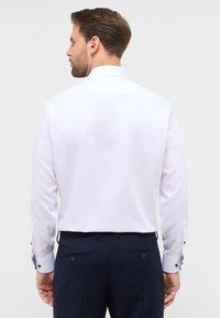 Weißes Langarmhemd mit klassischem Kragen, ausgestattet mit knöpfbaren Manschetten und glatter Textur. Getragen mit dunkelblauen Hosen.