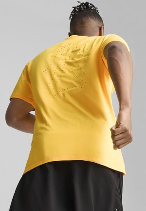 Camiseta atlética amarilla de mangas cortas, con detalles perforados en la parte de atrás, combinada con pantalones cortos negros para un look deportivo.