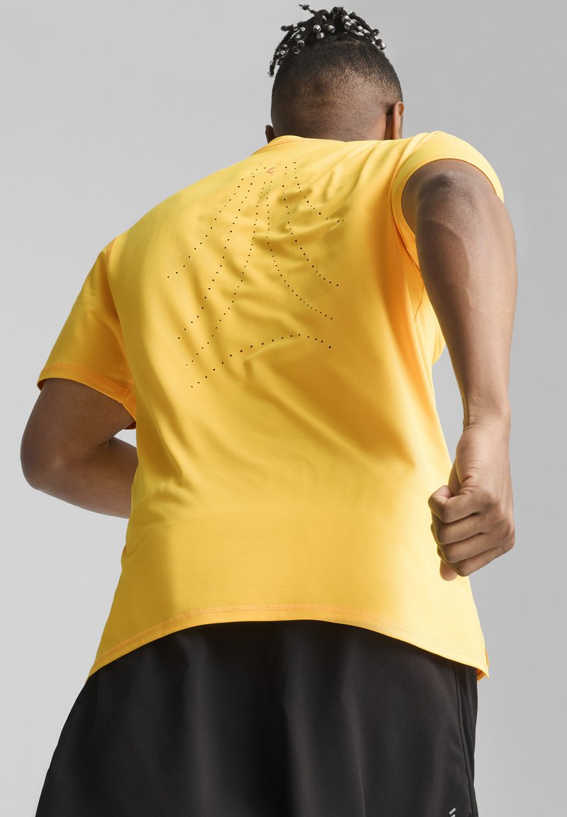 T-shirt atletico giallo con maniche corte, caratterizzato da dettagli perforati sulla schiena, abbinato a pantaloni corti neri per un look sportivo.
